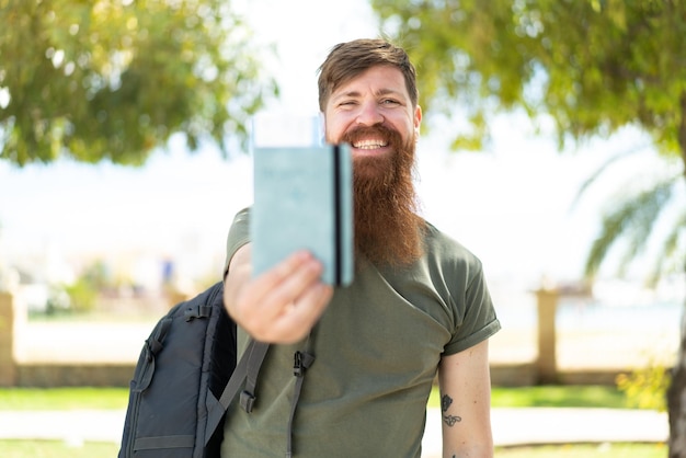 Homem ruivo com barba segurando um passaporte ao ar livre com expressão feliz