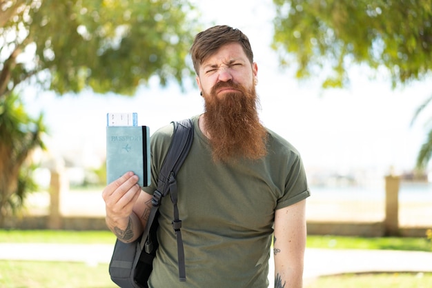 Homem ruivo com barba segurando um passaporte ao ar livre com expressão triste