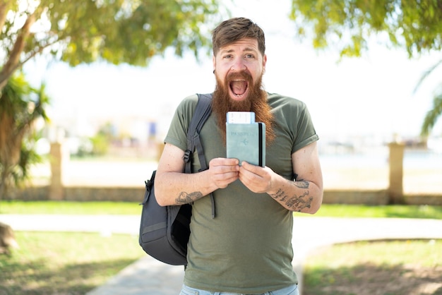 Homem ruivo com barba segurando um passaporte ao ar livre com surpresa e expressão facial chocada