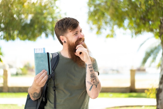 Homem ruivo com barba segurando um passaporte ao ar livre pensando em uma ideia e olhando de lado