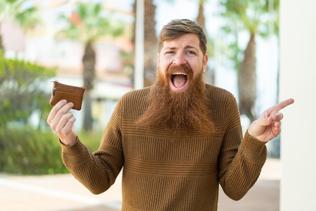 Homem ruivo com barba segurando uma carteira ao ar livre surpreso e apontando o dedo para o lado
