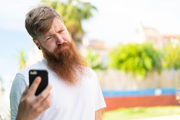Homem ruivo com barba usando telefone celular ao ar livre com expressão triste