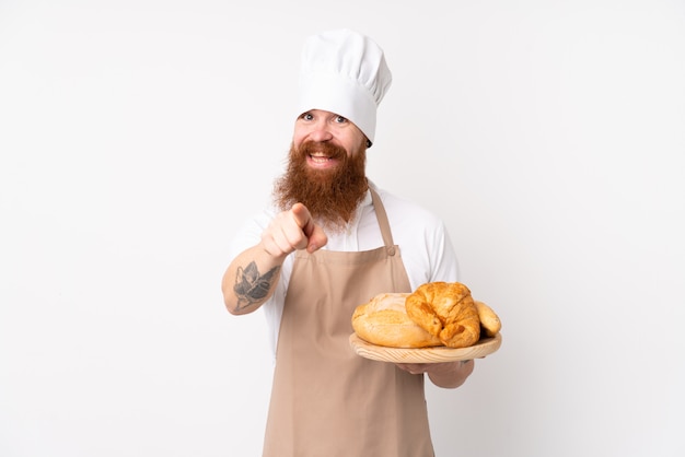 Homem ruivo de uniforme de chef. Padeiro masculino segurando uma mesa com vários pães aponta o dedo para você com uma expressão confiante
