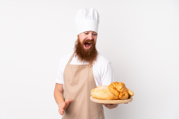 Homem ruivo de uniforme de chef. Padeiro masculino segurando uma mesa com vários pães com surpresa e expressão facial chocado