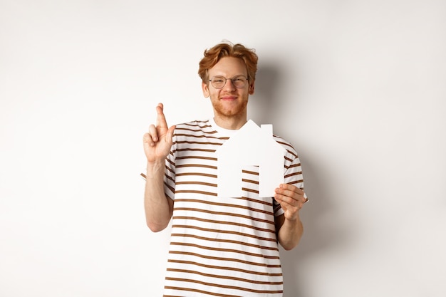 Homem ruivo esperançoso, sonhando em comprar uma casa, segurando o recorte de papel em casa e os dedos cruzados para dar sorte, fazendo um pedido, de pé sobre um fundo branco.