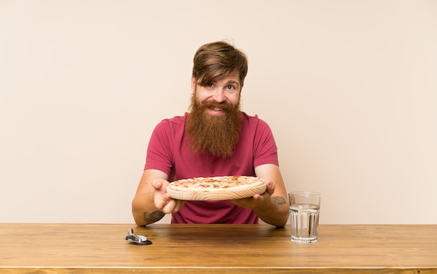 Homem ruivo feliz com barba longa em uma mesa e com uma pizza