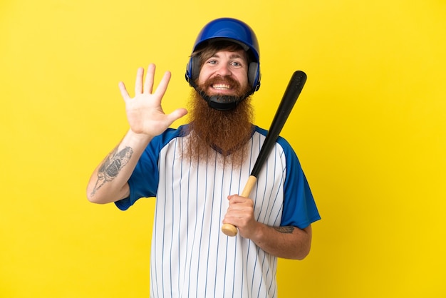Homem ruivo jogador de beisebol com capacete e taco isolado em um fundo amarelo contando cinco com os dedos
