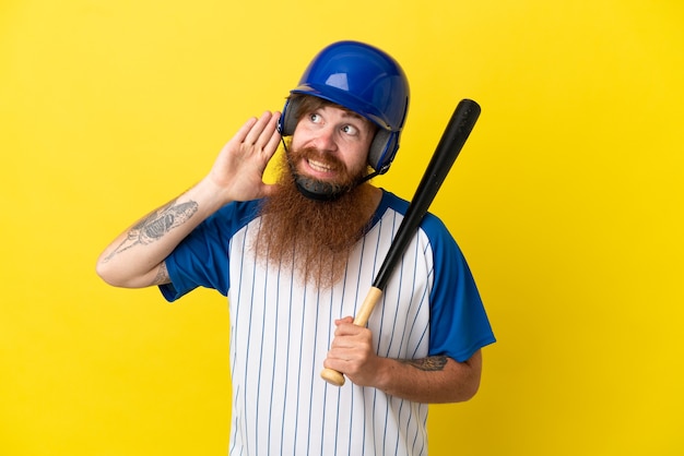 Homem ruivo jogador de beisebol com capacete e taco isolado em um fundo amarelo ouvindo algo colocando a mão na orelha