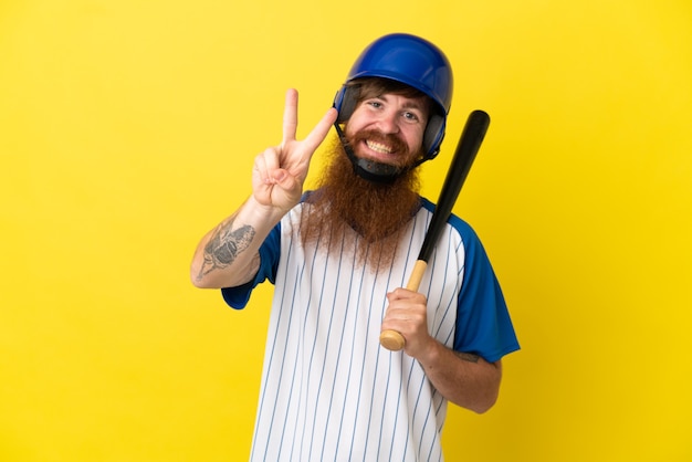 Homem ruivo jogador de beisebol com capacete e taco isolado em um fundo amarelo sorrindo e mostrando sinal de vitória