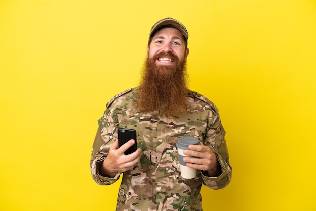 Homem ruivo militar isolado em fundo amarelo segurando café para levar e um celular