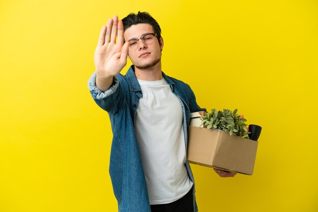 Homem russo fazendo um movimento enquanto apanha uma caixa cheia de coisas isoladas em um fundo amarelo fazendo um gesto de pare