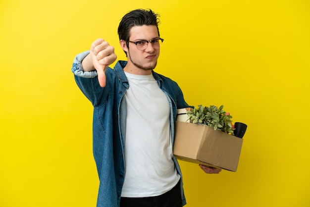 Homem russo se mexendo enquanto apanha uma caixa cheia de coisas isoladas na parede amarela mostrando o polegar para baixo com expressão negativa