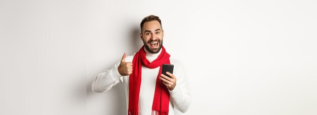 Foto homem satisfeito vendo algo bom online mostrando o polegar para cima enquanto usa o celular em pé sobre w