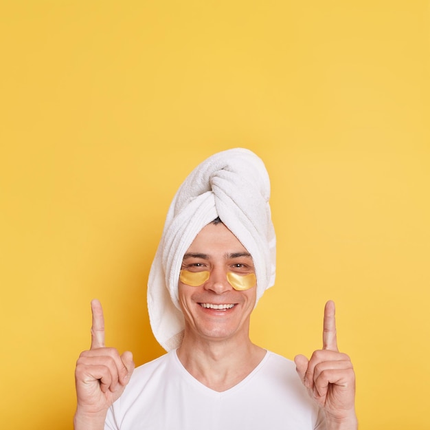 Homem satisfeito vestindo camiseta branca e toalha com manchas cosméticas sob os olhos apontando para cima com os dedos mostrando espaço para propaganda posando isolado sobre fundo amarelo