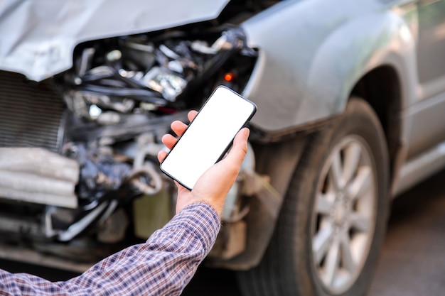 Homem segura a tela do telefone móvel branco da maquete nas mãos após um acidente de carro. Chamando o serviço de seguro no aplicativo da web para o local do acidente de carro. Smartphone na frente de um carro acidentado.