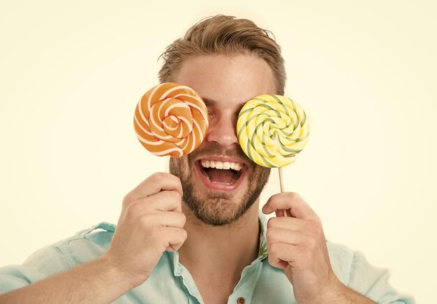 Homem segura grandes pirulitos nos olhos como óculos Conceito de guloso Cara no rosto sorridente segura dois pirulitos coloridos gigantes nas mãos isoladas no fundo branco Homem com cerdas gosta de doces