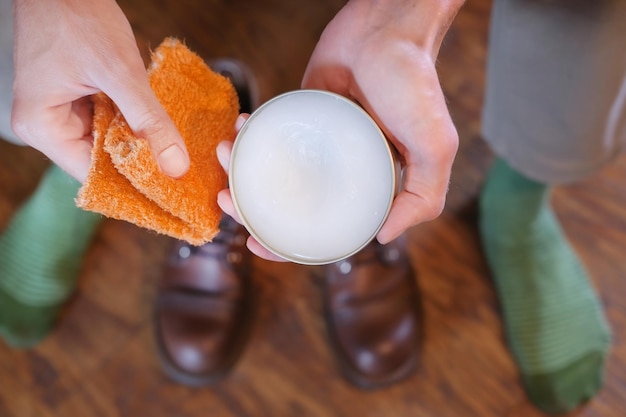 homem segura um pote de creme para sapatos e um guardanapo ao lado de botas de couro, limpando e cuidando de sapatos