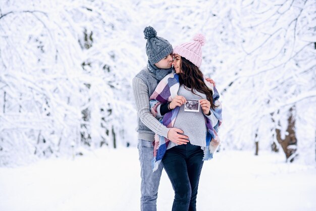Homem segurando a barriga grávida da esposa e a beija suavemente no nariz em um parque de inverno nevado