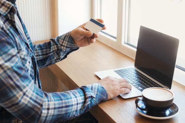 Homem segurando cartão de crédito bancário e fazendo pagamento online no internet banking, usando laptop e telefone