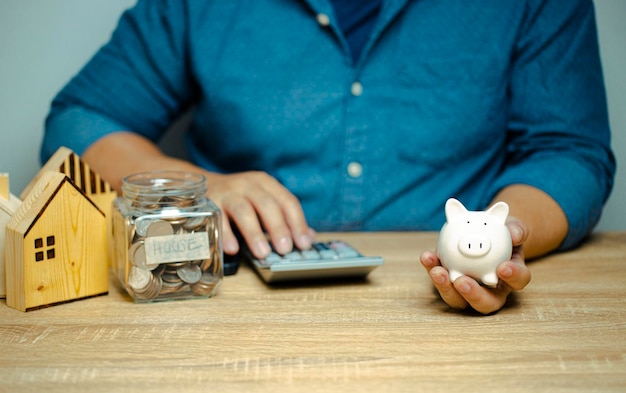 Homem segurando o cofrinho pressionando a calculadora Calcule receitas e despesas e economize para comprar uma casa