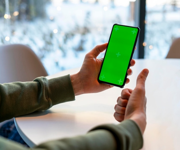 Homem segurando o smartphone com tela Chroma Key no café na mesa branca, mostrando os polegares para cima na tela verde