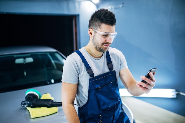 Foto homem segurando o telefone enquanto faz polimento de carro em sua oficina