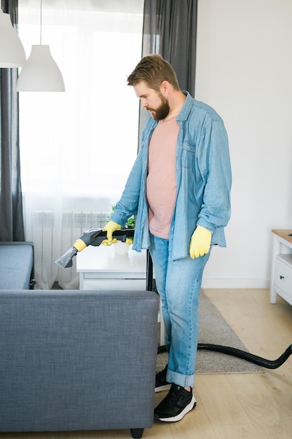 Homem segurando um aspirador de pó moderno e limpando sofá sujo com detergente profissional