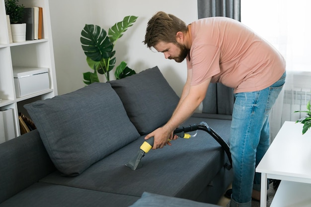 Homem segurando um aspirador de pó moderno e limpando sofá sujo com detergente profissional