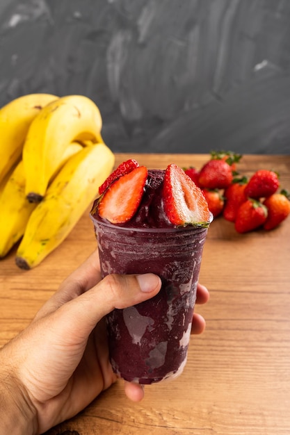 Homem segurando um Batido de Sorvete Brasileiro de Açaí Berry em copo plástico com Morangos. Sobre uma mesa de madeira e um fundo cinza de verão com frutas. Vista frontal para menu e mídia social