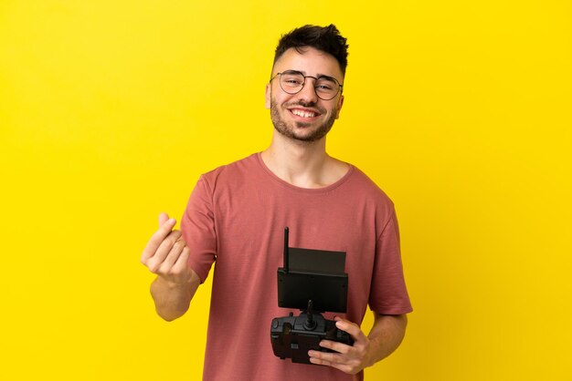 Homem segurando um controle remoto de drone isolado em um fundo amarelo fazendo gesto de dinheiro