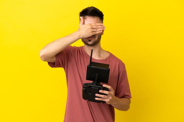 Homem segurando um controle remoto drone isolado em fundo amarelo, cobrindo os olhos com as mãos. Não quero ver nada