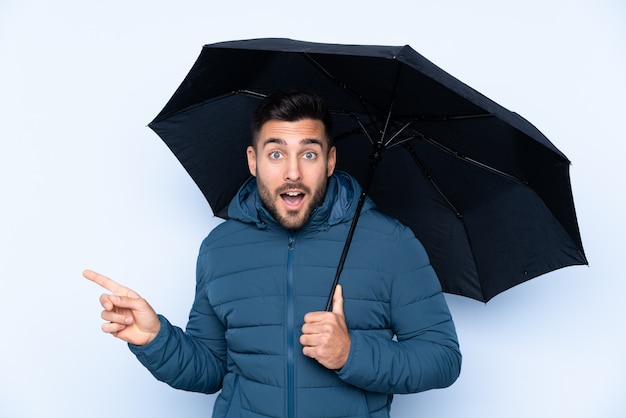 Homem segurando um guarda-chuva sobre parede isolada surpreendeu e apontando o dedo para o lado