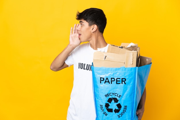 Homem segurando um saco de reciclagem cheio de papel para reciclar sobre amarelo isolado gritando com a boca aberta para o lado
