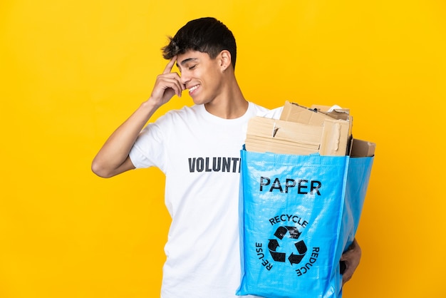 Homem segurando um saco de reciclagem cheio de papel para reciclar sobre amarelo rindo