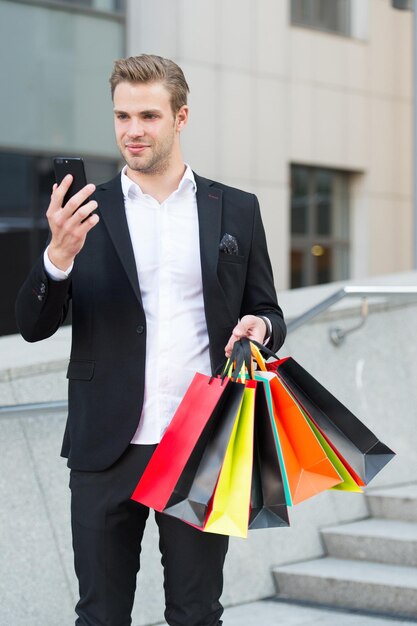 Homem segurar sacolas enquanto verifica o desconto online com o smartphone. Cara carrega sacolas de compras de monte. Ofertas lucrativas, compras na sexta-feira negra. Homem segura sacos de papel depois de fazer compras. Conceito de desconto de sexta-feira negra.