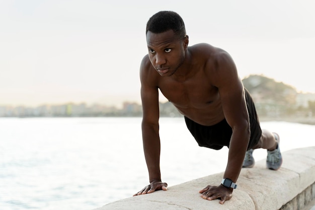 Homem sem camisa afro-americano fazendo flexões na praia com rosto concentrado