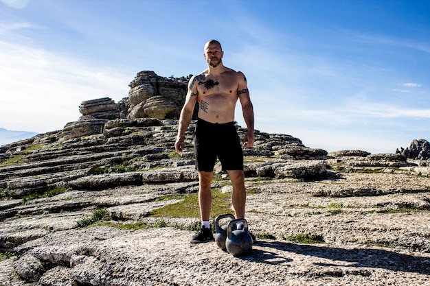 Homem sem camisa com dois kettlebells em cima da montanha