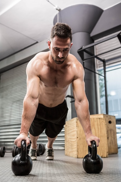 Homem sem camisa fazendo push-up com kettlebells no ginásio crossfit