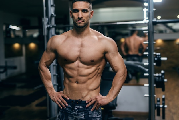 Homem sem camisa muscular com as mãos nos quadris, posando em uma academia