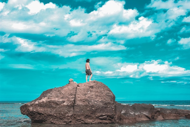 Homem sem camisa olhando para o mar enquanto está de pé na rocha contra o céu azul