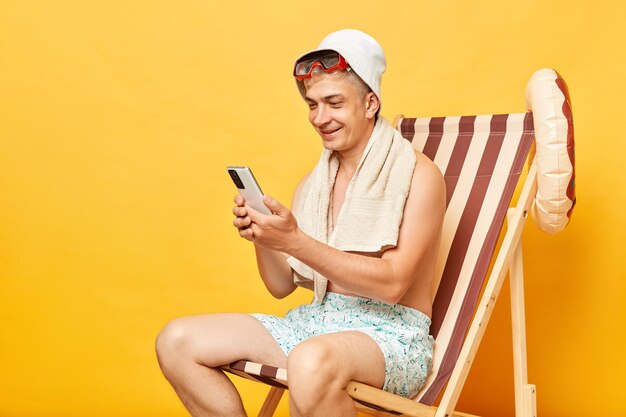 Homem sem camisa sorridente e feliz sentado em uma cadeira de terra isolado sobre um fundo amarelo descansando em um resort de praia usando telefone celular verificando redes sociais