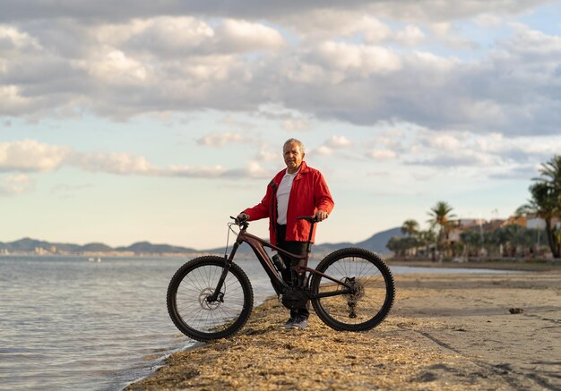 Foto homem sênior à beira-mar com uma bicicleta de montanha elétrica sorrindo olhando para a câmera ao pôr do sol