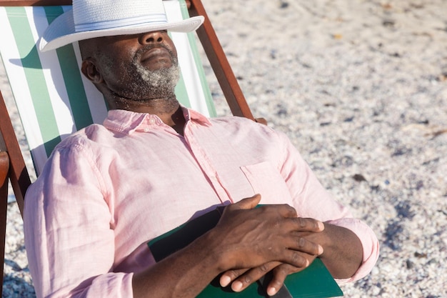 Homem sênior afro-americano aposentado dormindo com livro e chapéu sobre os olhos na cadeira dobrável na praia