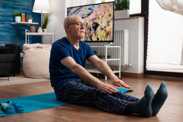 Homem sênior alongando os músculos das pernas enquanto está sentado em um tapete de ioga na sala de estar durante um treino de pilates