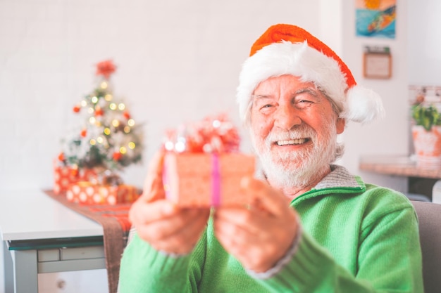 Homem sênior aposentado com chapéu de papai noel e roupas quentes comemorando o natal em casa papai noel velho sorrindo enquanto segura o presente de natal homem idoso segurando a caixa de presente de natalxa