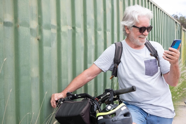 Homem sênior ativo sorridente usando telefone celular enquanto desfruta de atividade esportiva com bicicleta elétrica no conceito de mobilidade sustentável ao ar livre