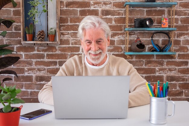 Homem sênior atraente relaxado usando o laptop em casa. Parede de tijolos no fundo