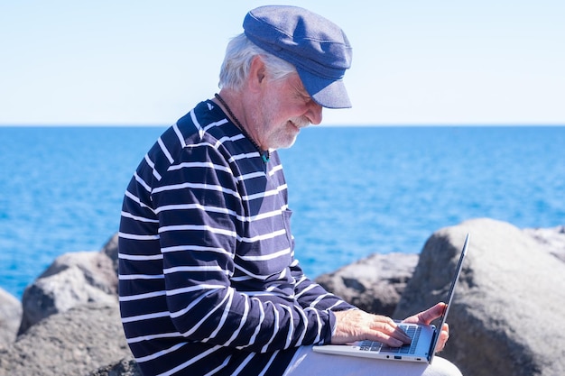 Homem sênior barbudo digitando no laptop enquanto está sentado perto do mar em trabalho remoto