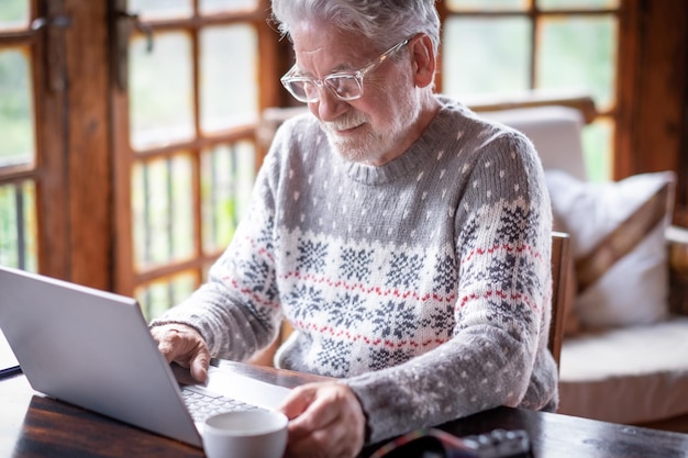 Homem sênior barbudo sorridente vestindo suéter sentado na sala de estar usando computador portátil segurando uma xícara de café Homem idoso despreocupado trabalhando em casa desfrutando de tecnologia e social