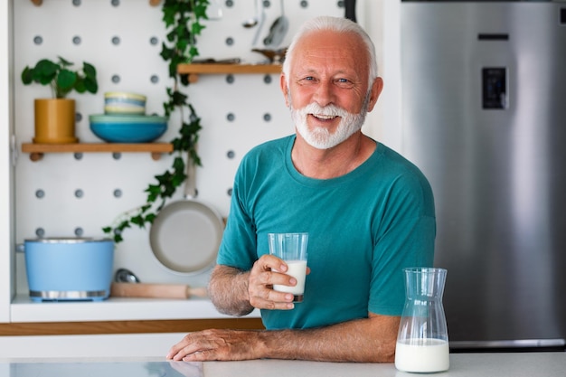 Homem sênior bebendo um copo de leite com uma cara feliz em pé e sorrindo Homem sênior bonito bebendo um copo de leite fresco na cozinha